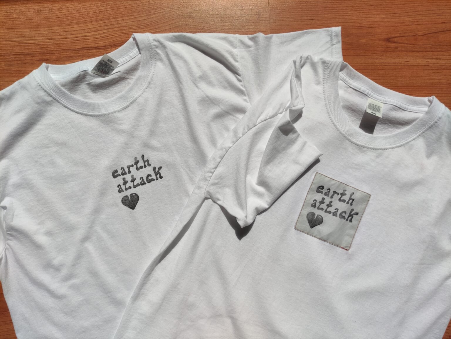 Photo cliquable menant à la page de présentation du projet "Earth Attack" - La photo montre deux t-shirt blancs superposés, sur lesquels ont voit le logo, positionné au niveau du centre de la poitrine. Sur le t-shirt de gauche, le logo est imprimé à même le t-shirt tandis que sur celui de droite, il est imprimé sur un tissus carré légèrement plus foncé et la pièce est cousue sur le t-shirt avec un fil rouge.